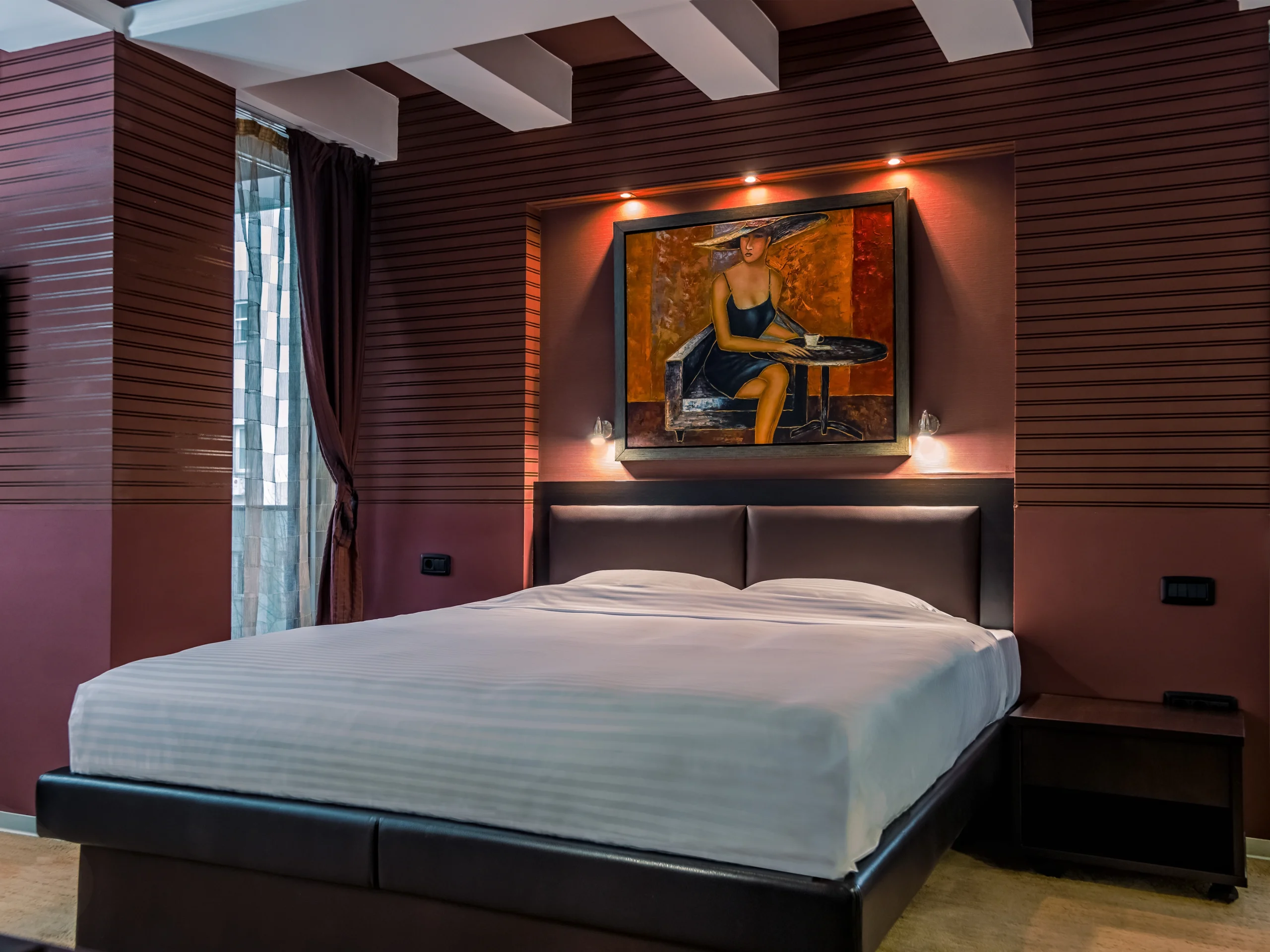 A modern bedroom at Sienna Apartments Skopje features a neatly made bed, dark brown walls, a painting of a seated person above the bed, side tables, and a window with sheer curtains.